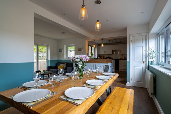 A dining area with a long wooden table set for six, a vase with flowers, and pendant lighting.