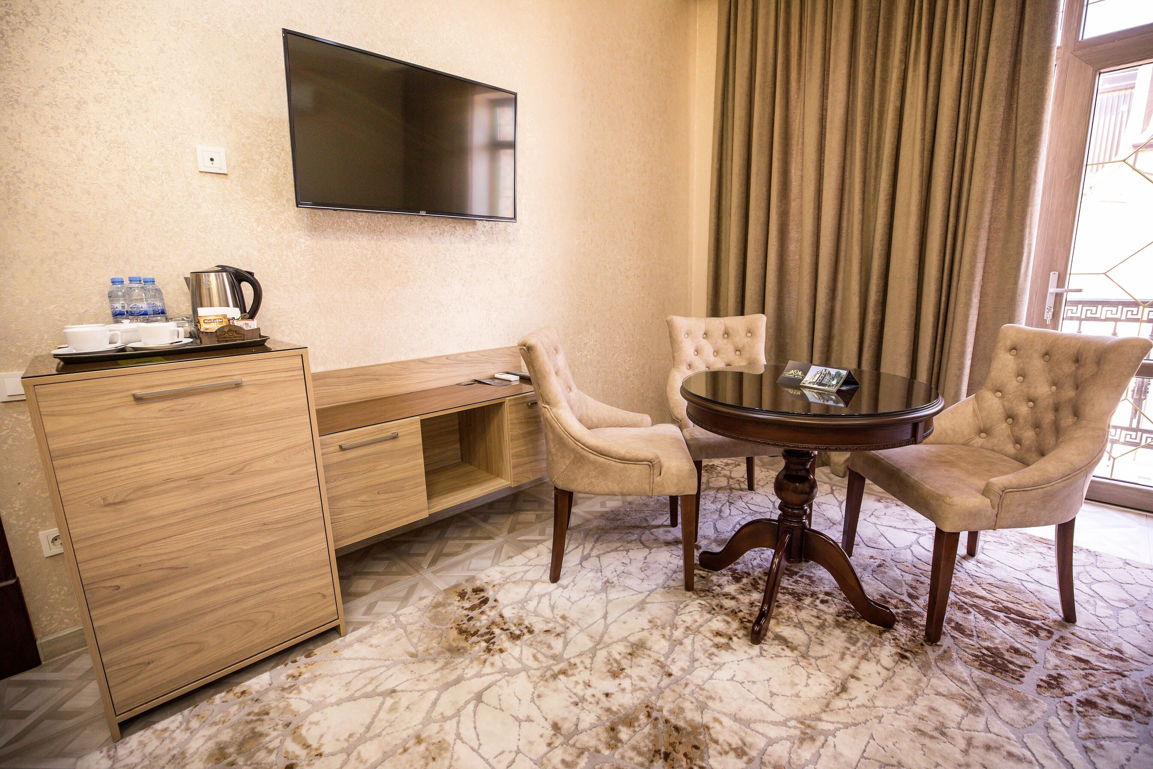 A hotel room with a wooden desk, two armchairs, a round table, a flat-screen TV, and a large window with curtains.