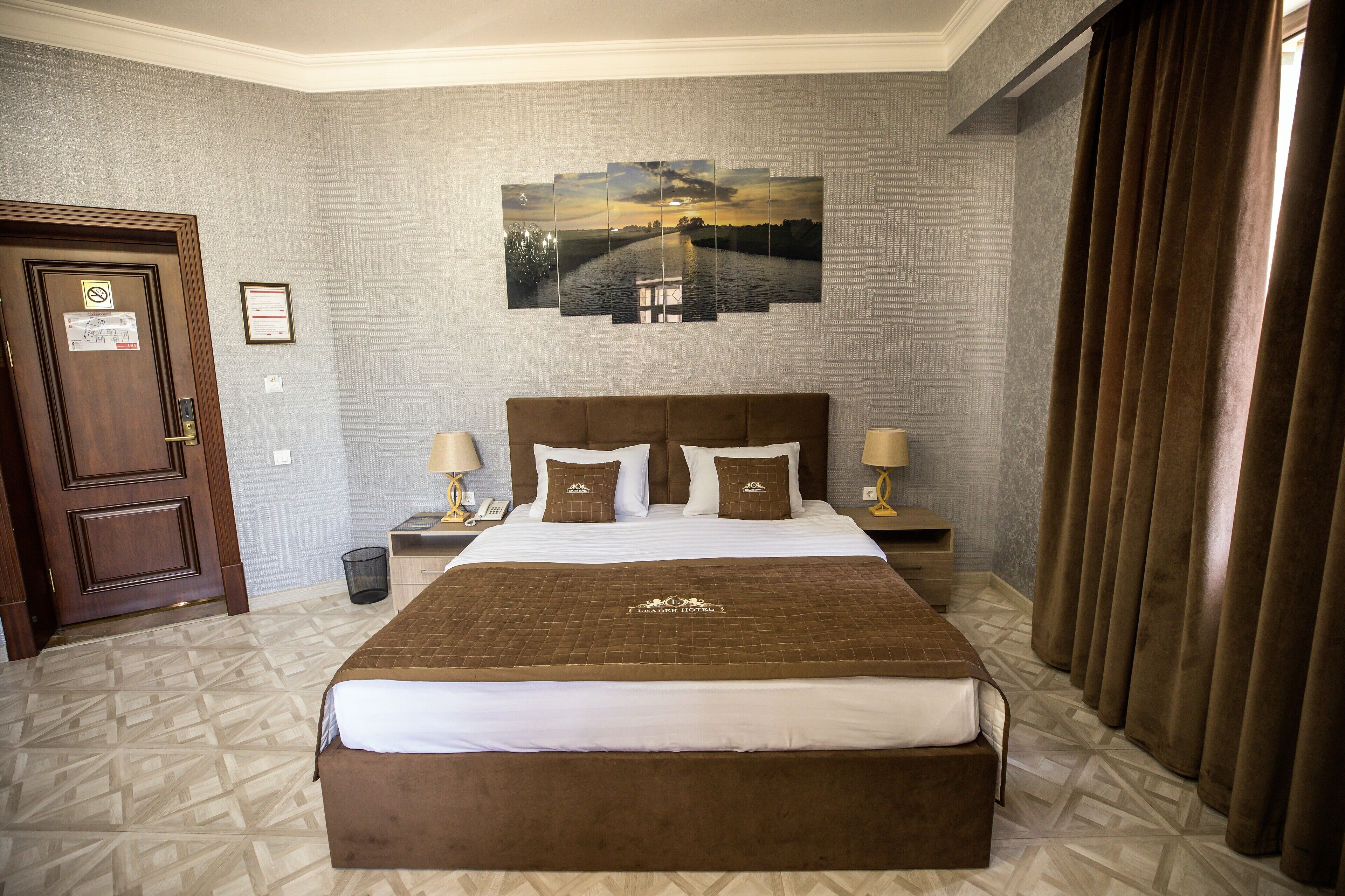Habitación de hotel con una cama grande, dos mesitas de noche, una lámpara y un cuadro en la pared, además de una puerta con un cartel.