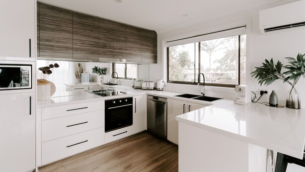 A modern kitchen with white cabinetry, a built-in oven, a dishwasher, and a large window providing natural light.