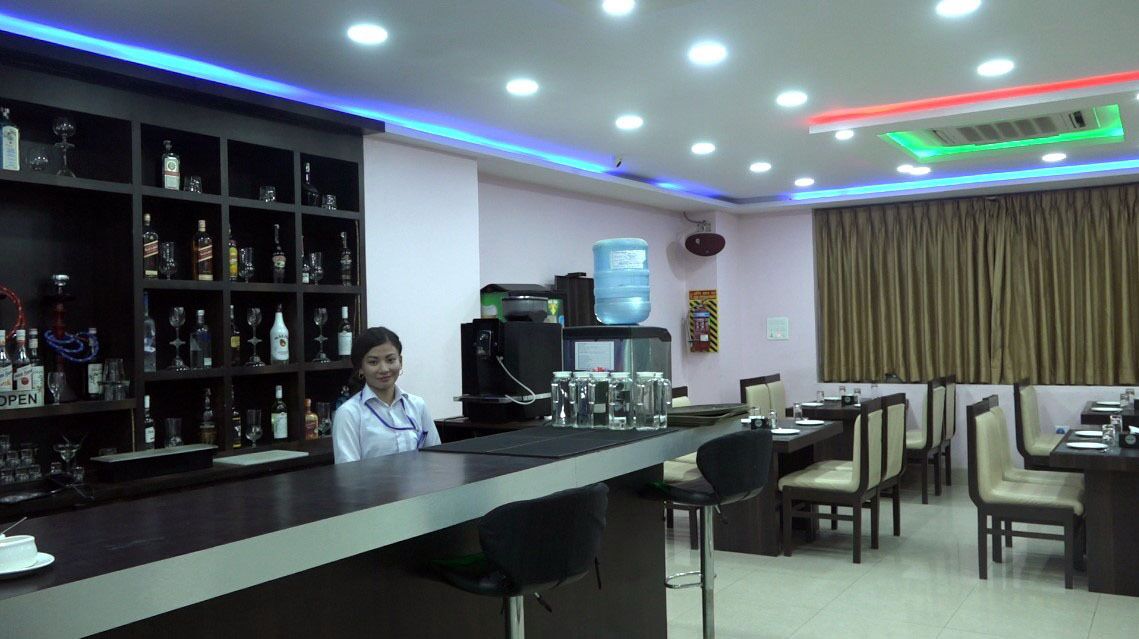 Zona de bar con estantes llenos de botellas de licores, un dispensador de agua y una máquina de café. Hay mesas y sillas dispuestas para comer.