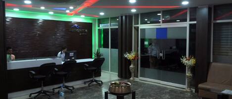 A reception area with a modern design, featuring a reception desk, black office chairs, a small table with a vase of flowers, and a brown leather couch.