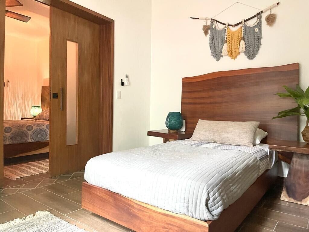 A bedroom with a wooden bed, a nightstand, a wall-mounted lamp, and a plant.