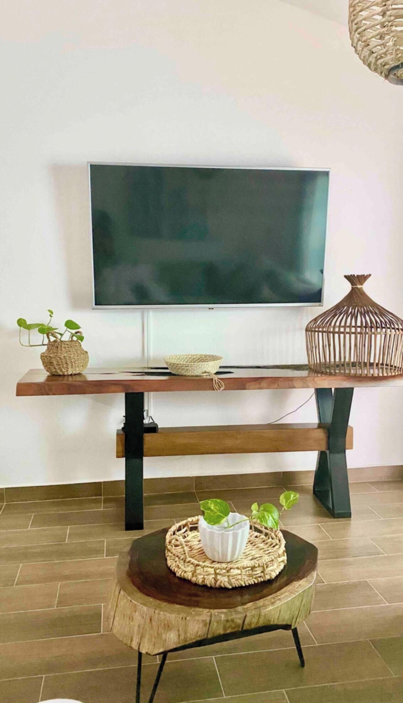 Un salón moderno con un televisor de pantalla plana montado en la pared, una mesa de centro de madera con una cesta tejida, y una pequeña mesa redonda con una planta.