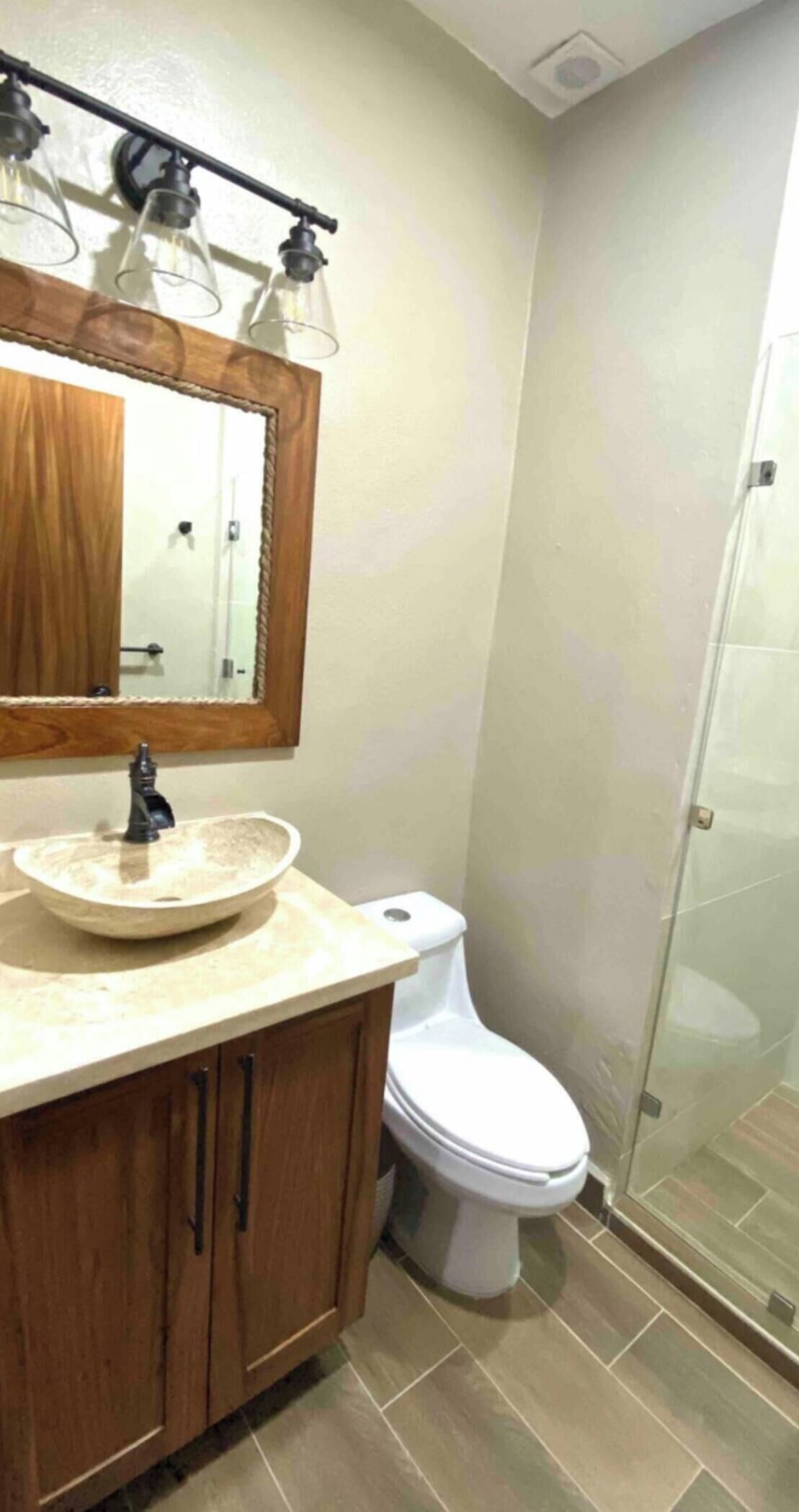 Un baño con tocador de madera, lavamanos redondo, inodoro blanco y cabina de ducha de vidrio.