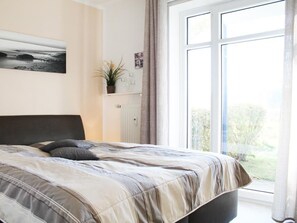 A bedroom with a bed, a window, a plant, and a painting on the wall.