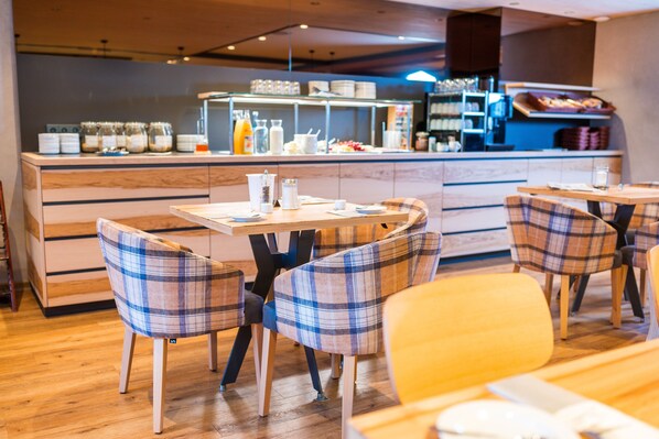 Un café avec des tables en bois et des chaises à carreaux, un comptoir où sont exposés des plats et des boissons, et une cuisine moderne en arrière-plan.