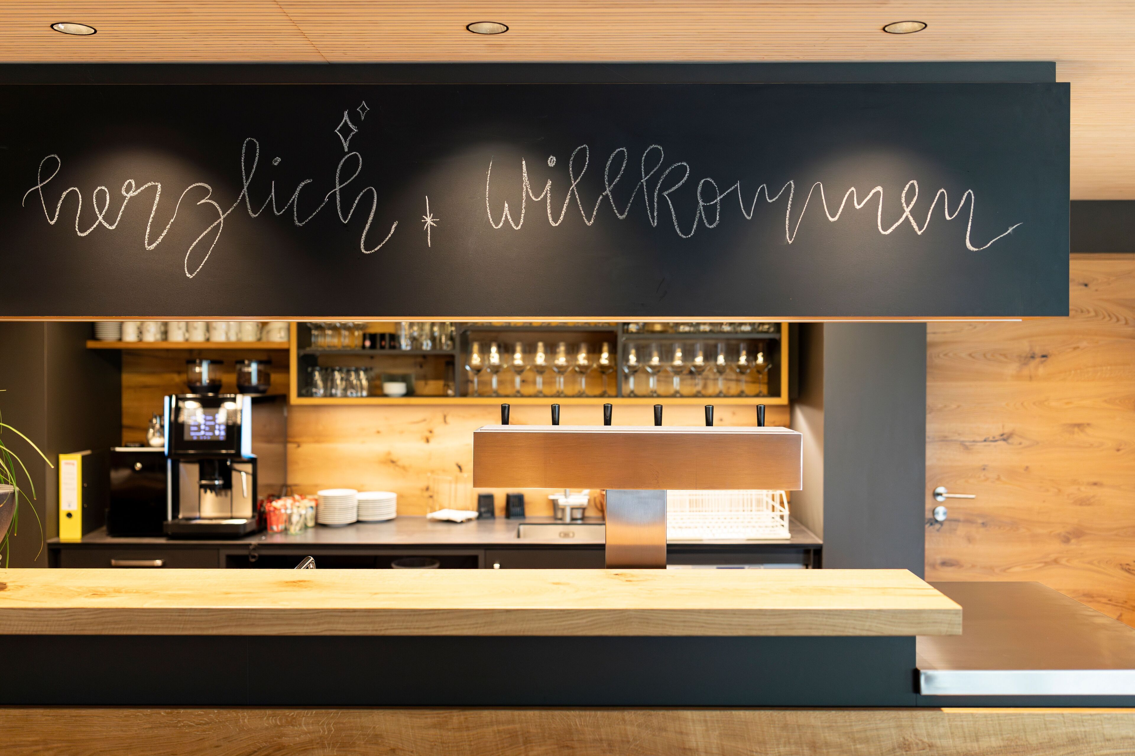 Een moderne bar met een krijtbord waarop staat geschreven 'herzlich Willkommen' en een koffieapparaat.