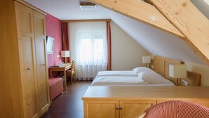 A hotel room with a wooden wardrobe, a desk with a chair, a bed with two pillows, and a window with curtains.