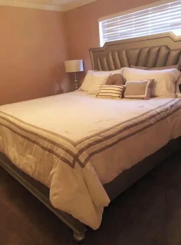 A neatly made bed with a headboard, a bedside lamp, and a window with blinds.