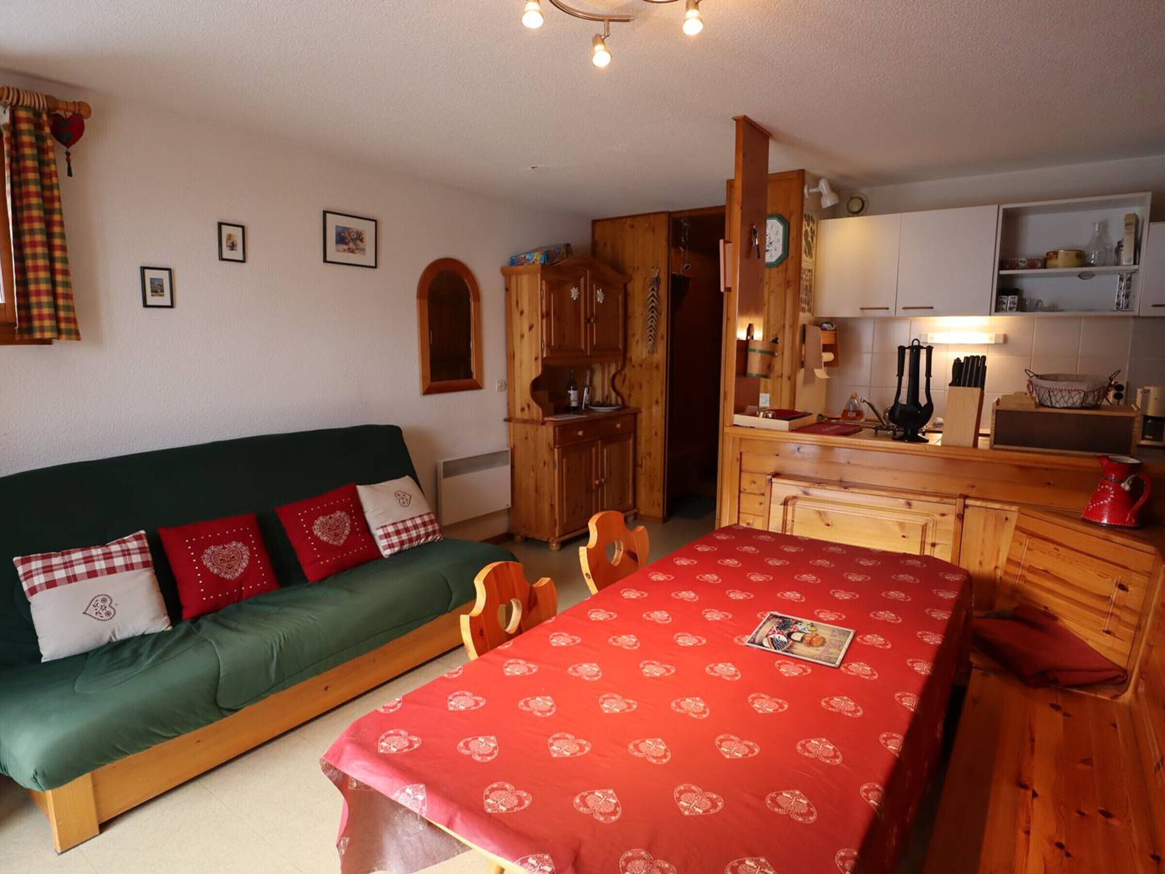A cozy room with a green sofa, a red table with a heart pattern, wooden cabinets, and a kitchen area.