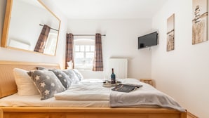 A neatly made bed with a wooden frame, a bedside table with a bottle and glasses, a TV mounted on the wall, and a window with curtains. - Villa Annabelle Fewo 7 Auszeit - Apartment 7 Auszeit (Binz)