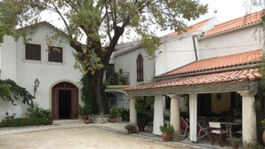 A courtyard with a large tree, a covered patio area with outdoor furniture, and a building with a tiled roof and arched doorway. - Casa dos Arrábidos - Turismo em Espaço Rural (Torres Novas)