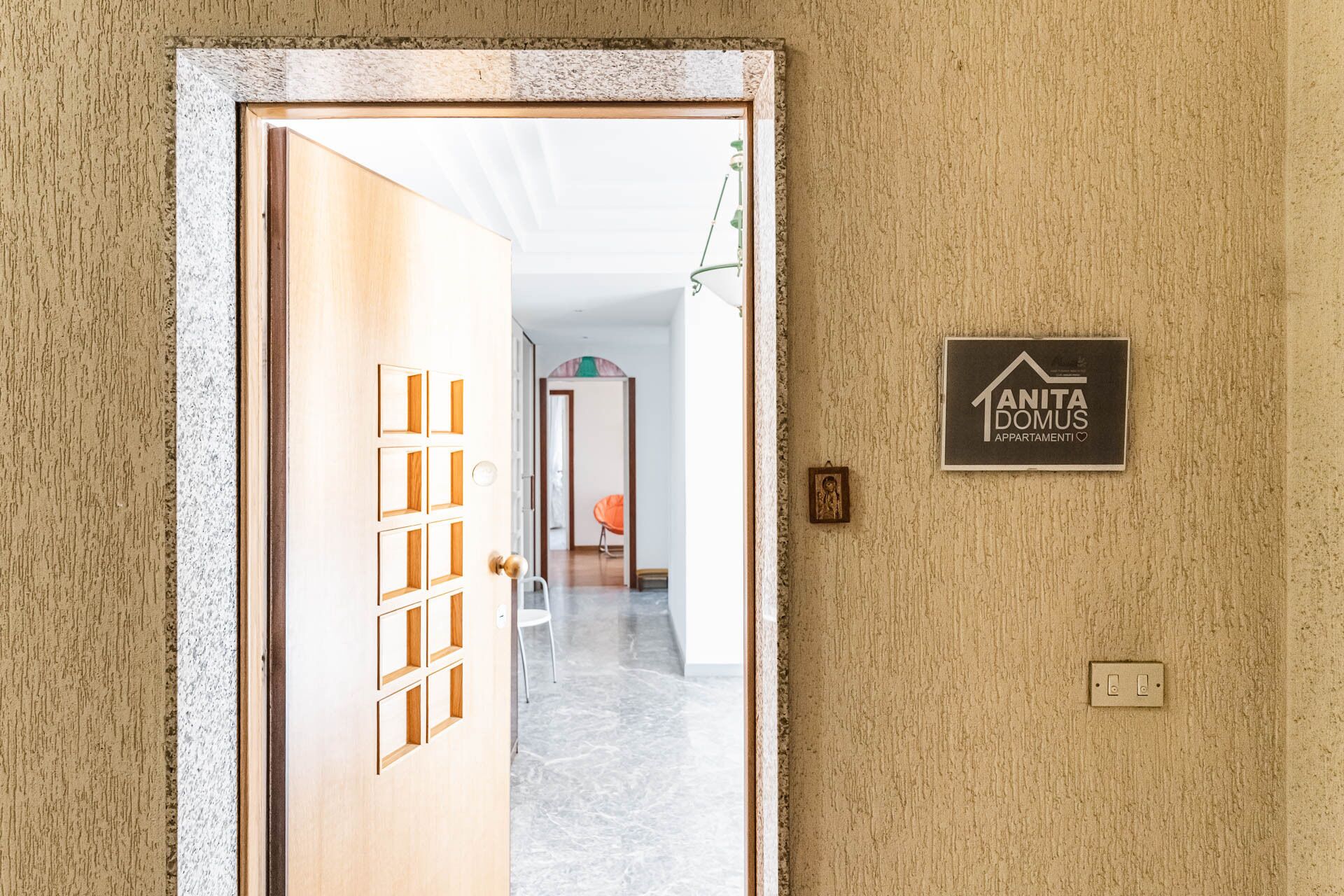 A hallway with a door leading to a room, a sign reading "ANITA DOMUS APARTAMENTI", and a light switch on the wall.