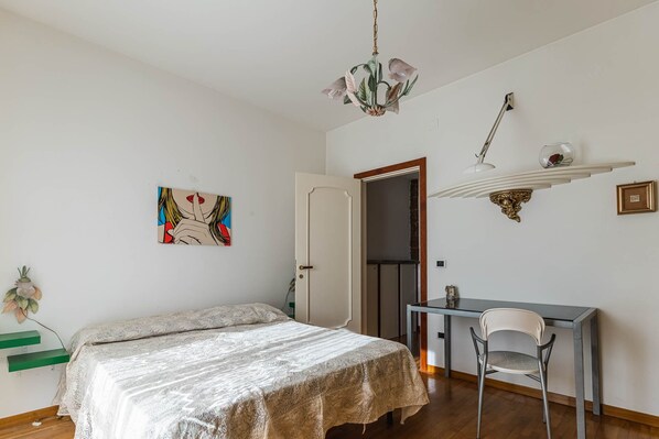 A bedroom with a bed, a desk, a chair, a hanging light fixture, and a door.