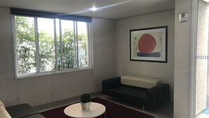 A modern living room with a large window, a round coffee table with a plant, a black sofa, and a framed abstract painting on the wall. - Apto Conforto C/ Netflix Amazon Prime Disponível (SP)