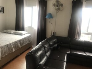 Ein Zimmer mit einem Bett, einer schwarzen Ledersofa, einem Standventilator und einer Lampe.