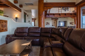 A spacious living room with a large leather sectional sofa, wooden coffee table, and a high wooden ceiling with exposed beams.