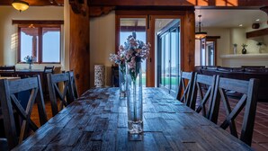 A rustic wooden dining table with chairs, a vase with flowers, and a view of the outdoors through large sliding doors.