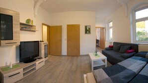 Ein modernes Wohnzimmer mit Flachbildfernseher, Holzmöbeln, einer Couch und einem Fenster, das für natürliches Licht sorgt.