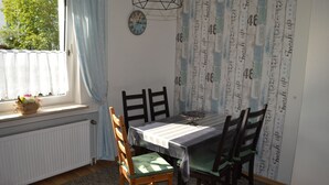 Un coin repas comprenant une table, des chaises, une fenêtre avec des rideaux, un radiateur et un mur recouvert d’un papier peint à motifs.