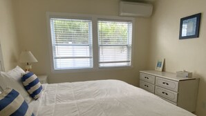 Una camera da letto con un letto grande, due finestre con tende, un comodino con una lampada, una cassettiera e un quadro incorniciato appeso al muro.