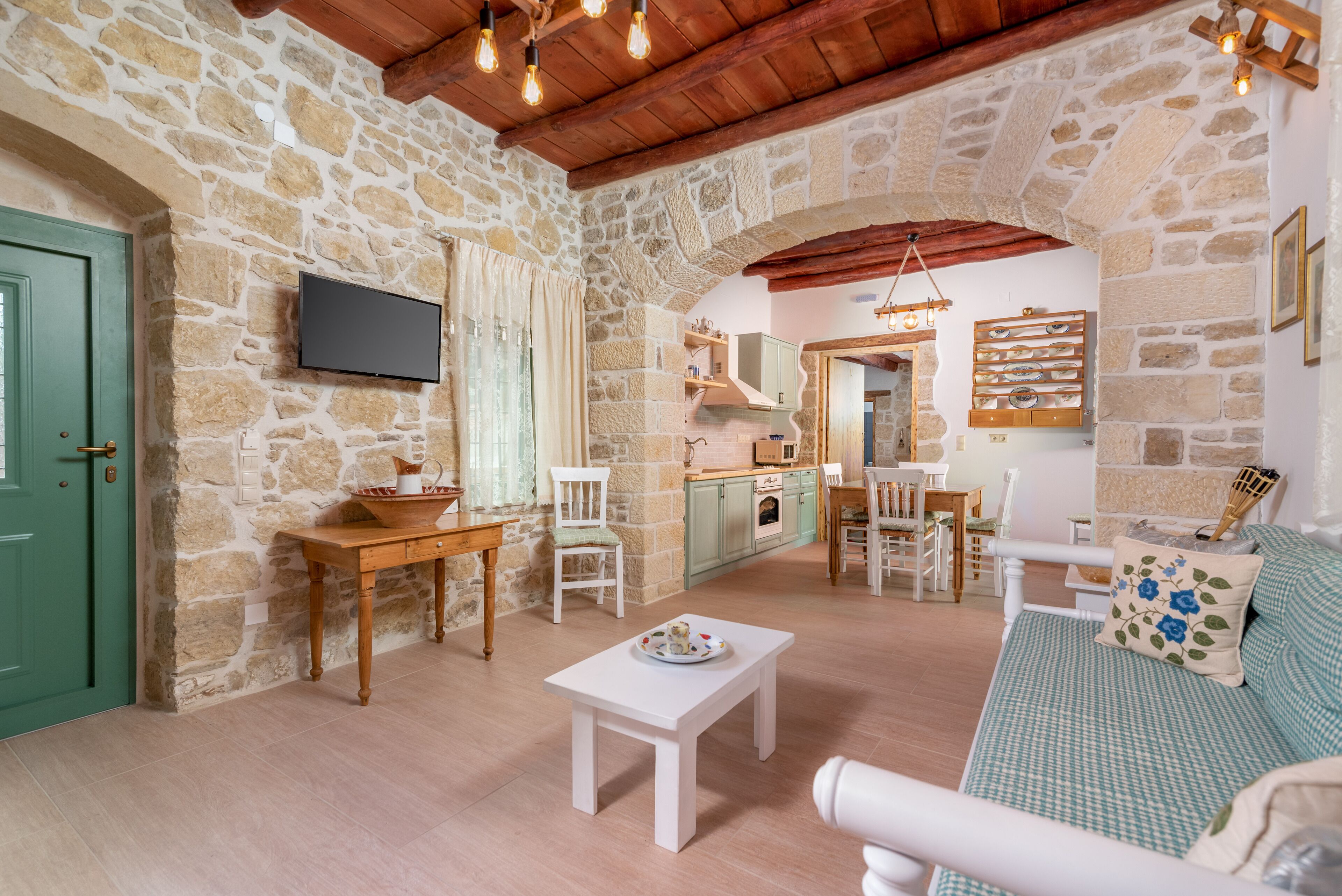 A cozy living room with a stone wall, wooden furniture, a television, and a kitchen in the background.