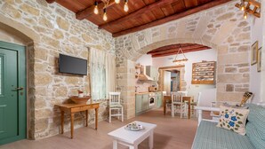 A cozy living room with a stone wall, wooden furniture, a television, and a kitchen in the background. - Joakim's villa  (herakleion)