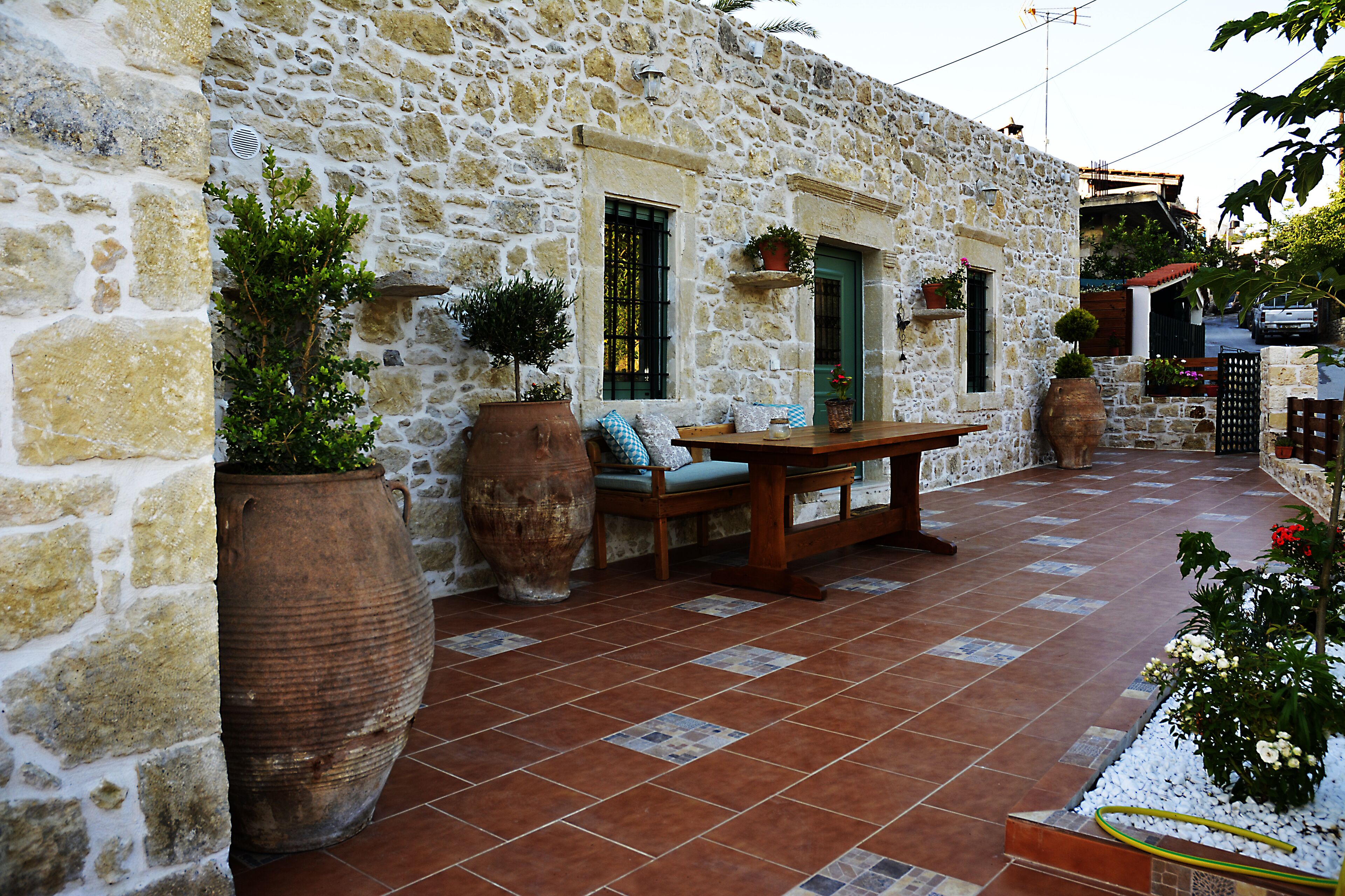 A stone building with a patio area featuring a wooden table and chairs, large planters, and a tiled floor.