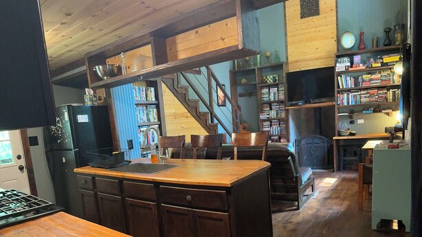 A kitchen with wooden cabinets, a central island, and a dining area with chairs. There is a staircase leading to an upper level.