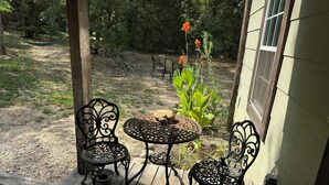 An outdoor patio with two ornate metal chairs and a round table, surrounded by a wooden deck and a garden with plants and flowers.