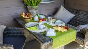 Ein Frühstückstisch auf einer Veranda mit Korbmöbeln, Tellern, Tassen und einem Brotkorb.