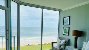 Habitación con un gran ventanal corredizo que da a una vista de la playa. Hay una silla de mimbre con cojín, una lámpara y dos cuadros en la pared.
