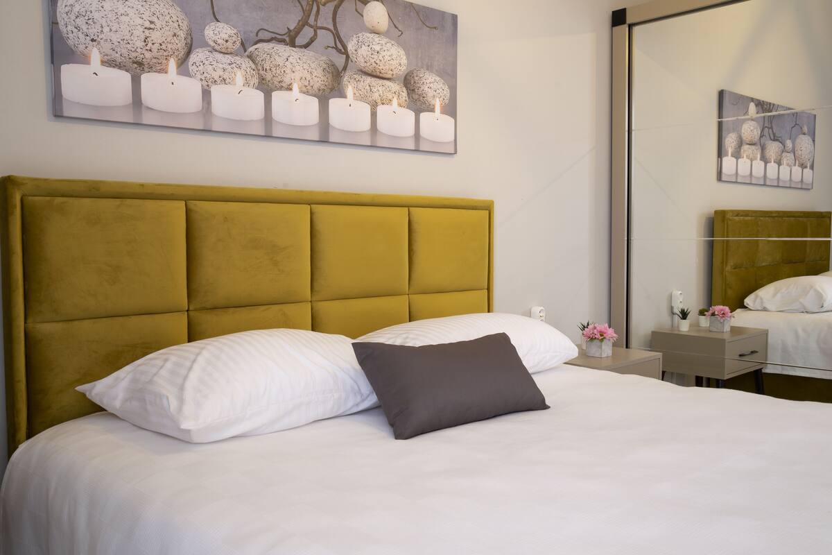 A neatly made bed with white and gray bedding, a mustard-colored headboard, and a painting of eggs on the wall above.