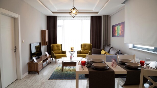A modern living room with a sofa, two armchairs, a coffee table, and a dining area with a table set for two. - Hatton Suites Esenboga (Ankara)