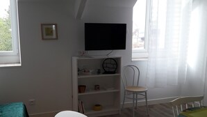 Une pièce avec un téléviseur fixé au mur, une chaise blanche, une petite table et une table à manger recouverte d’une nappe verte.
