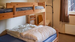 Ein Stockbett mit einem unterem Bett und einem Einzelbett darunter. Es gibt ein hölzernes Kopfteil, einen Nachttisch mit einem Stuhl und ein Fenster mit Vorhängen.