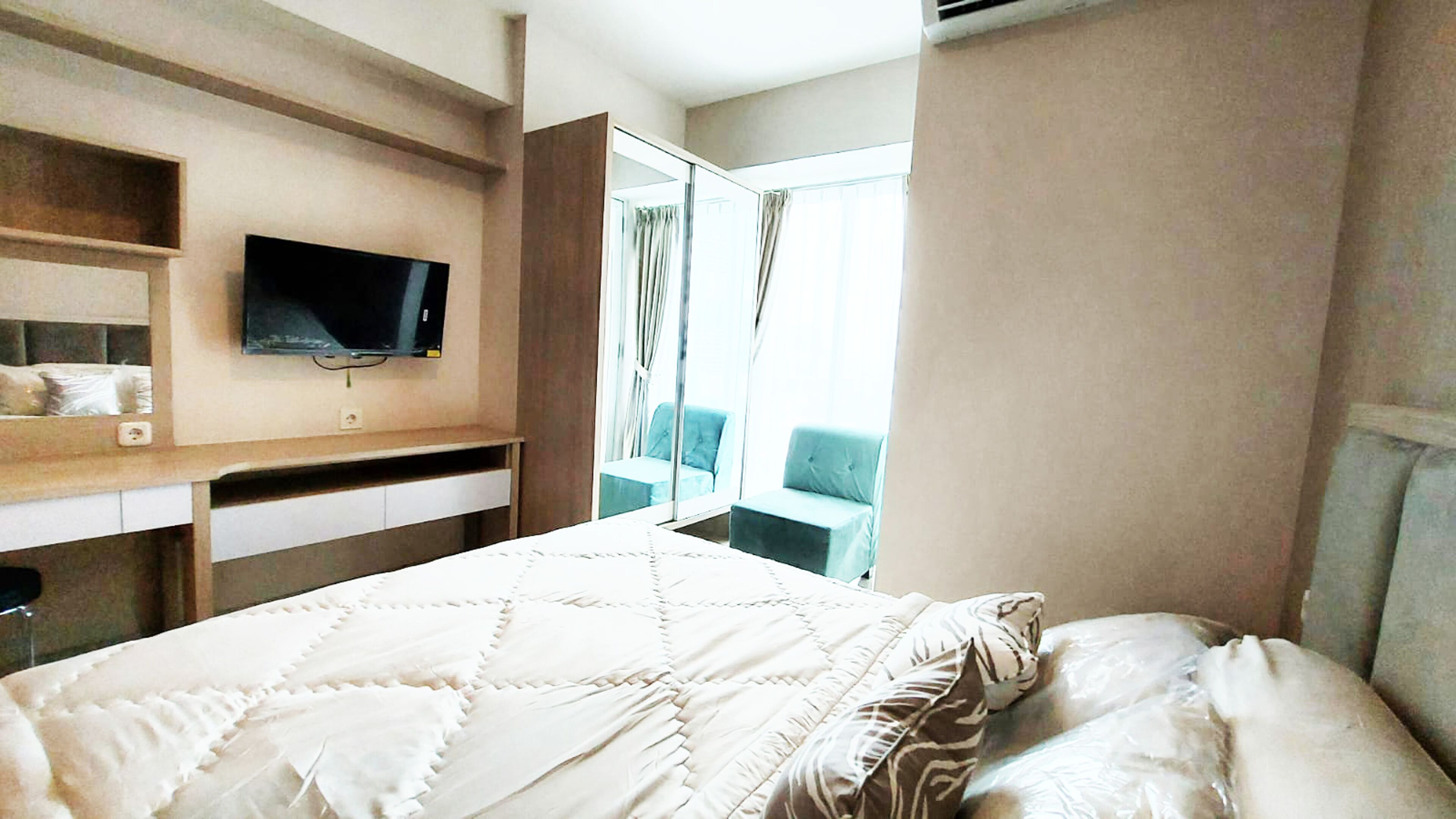 Ein Hotelzimmer mit Bett, Wandfernseher, Fenster mit Vorhängen und einer kleinen Sitzecke mit einem Sessel.