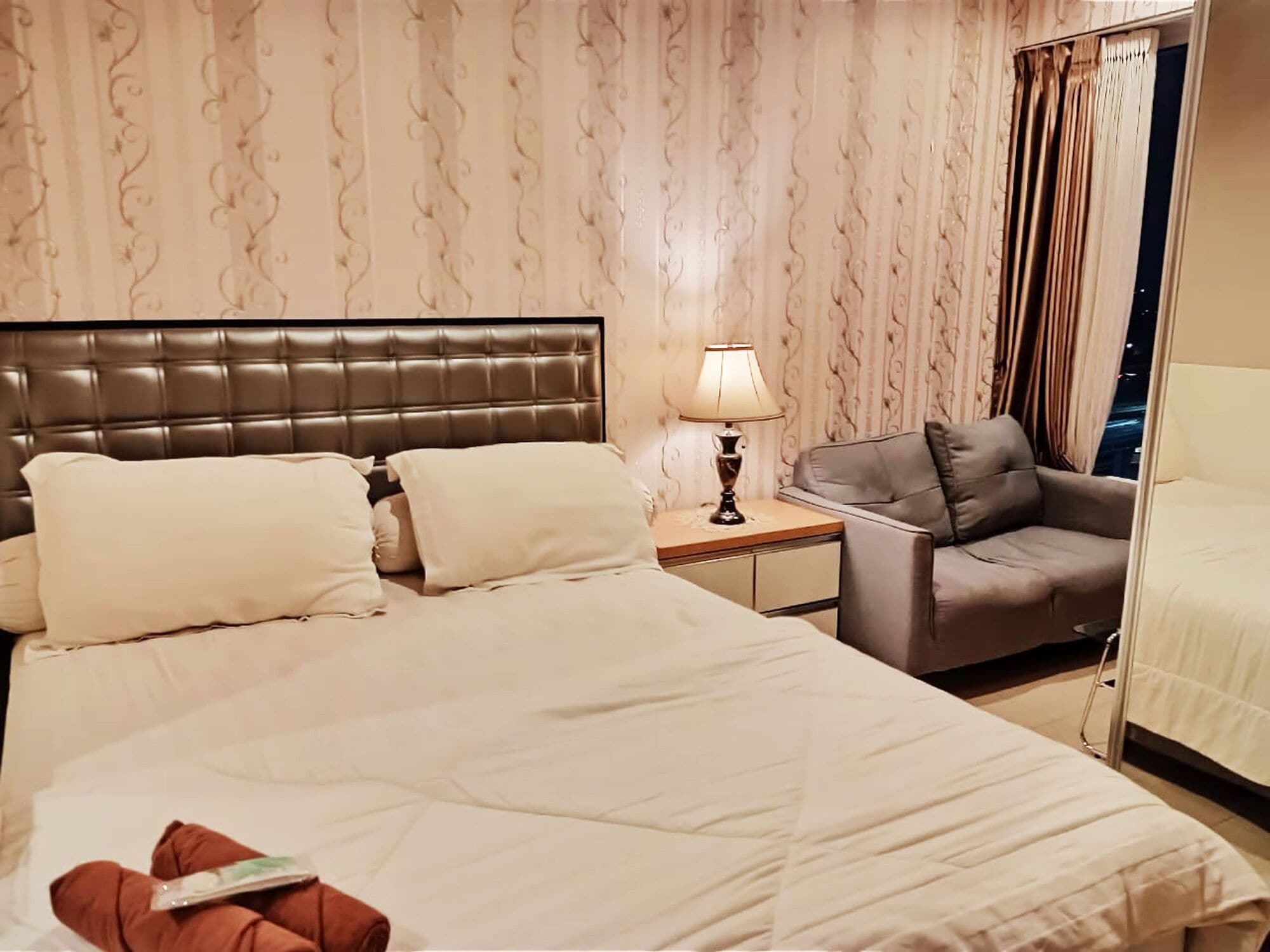 A hotel room with a large bed, a bedside table with a lamp, a grey armchair, and a window with curtains.