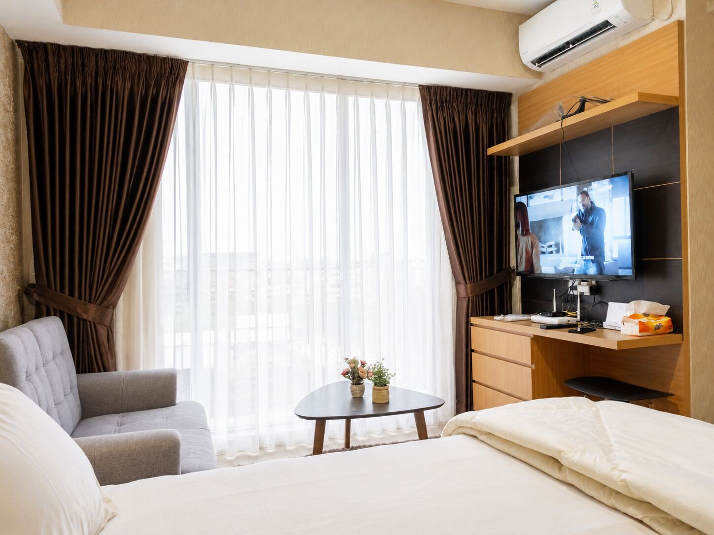 Quarto de hotel com cama, escrivaninha, televisão e janela com cortinas.