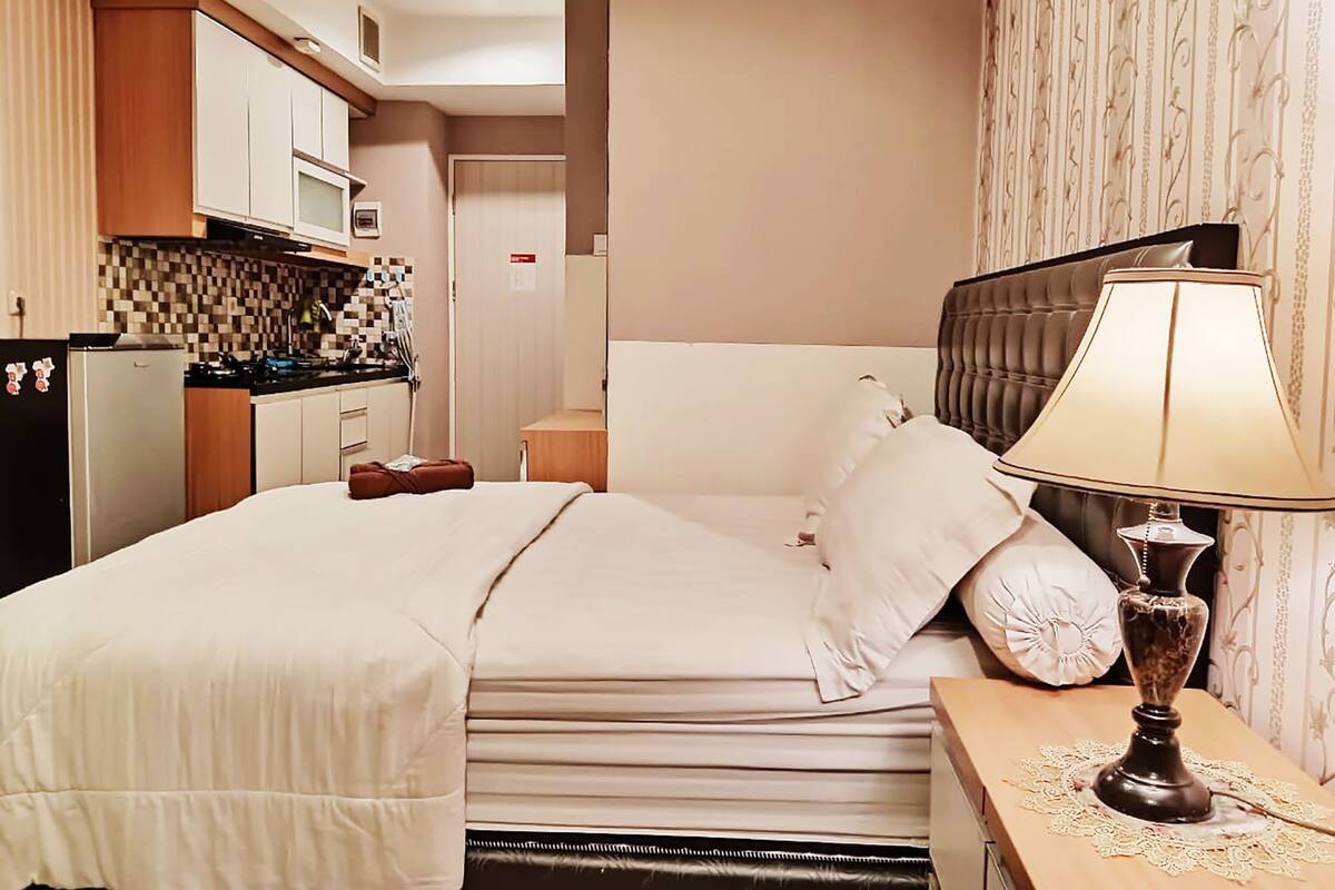 Ett hotellrum med en säng, ett sängbord med en lampa, en köksdel med skåp och ett kylskåp samt en badrumsdörr.