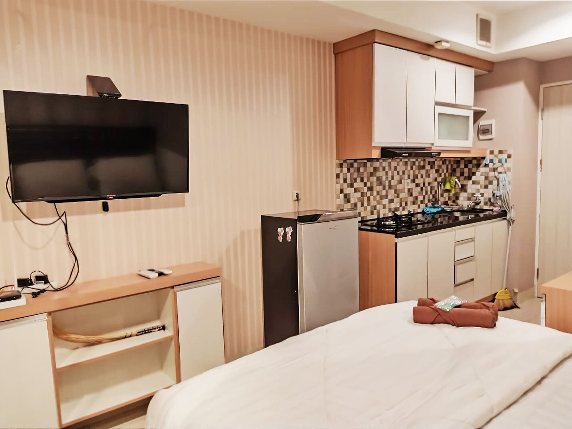 A compact hotel room with a kitchenette, a flat-screen TV, a bed with pillows, and a small shelf.