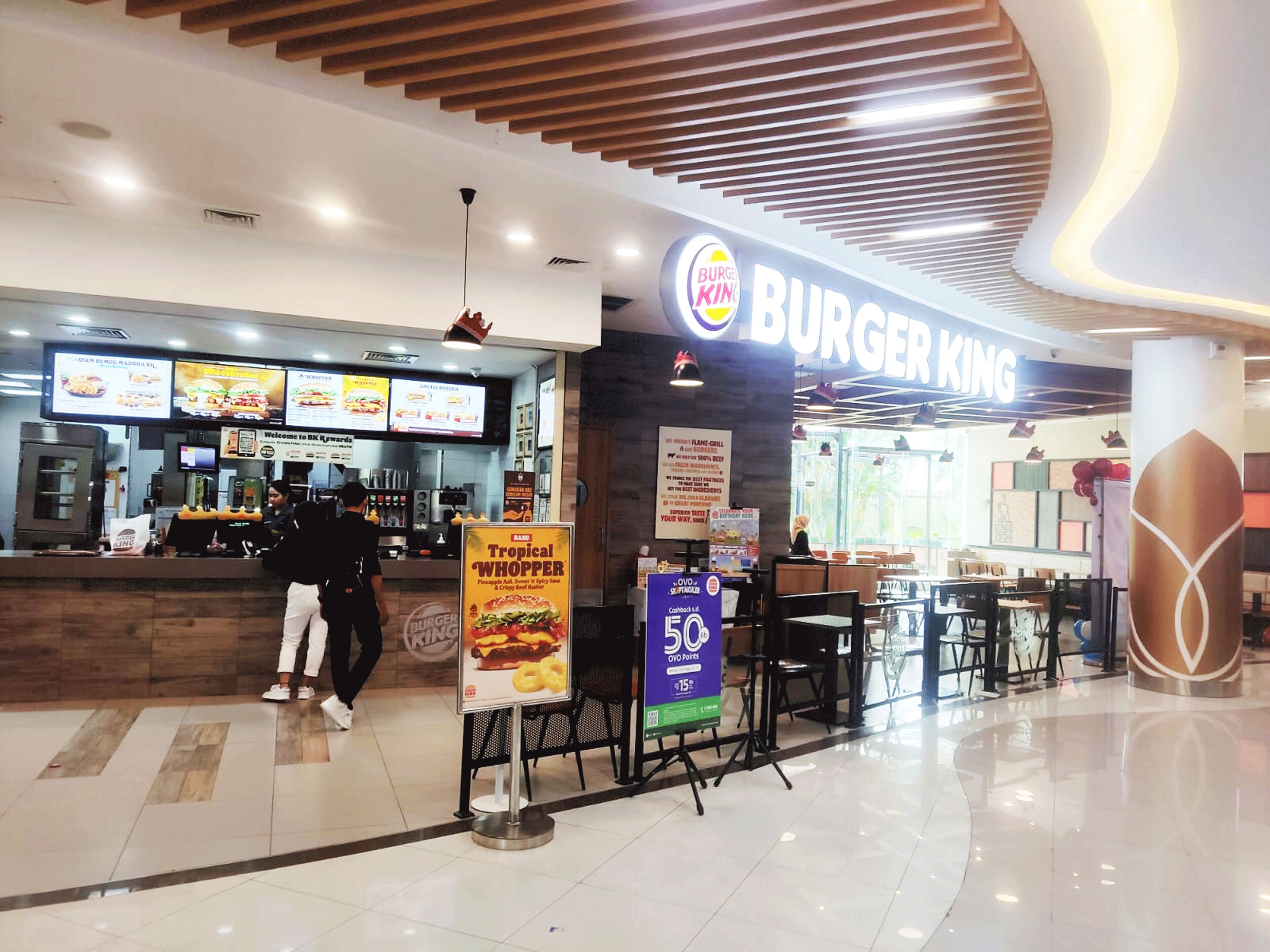 A Burger King restaurant with a modern interior, a digital menu board, and a promotional sign for the Tropical Whopper.