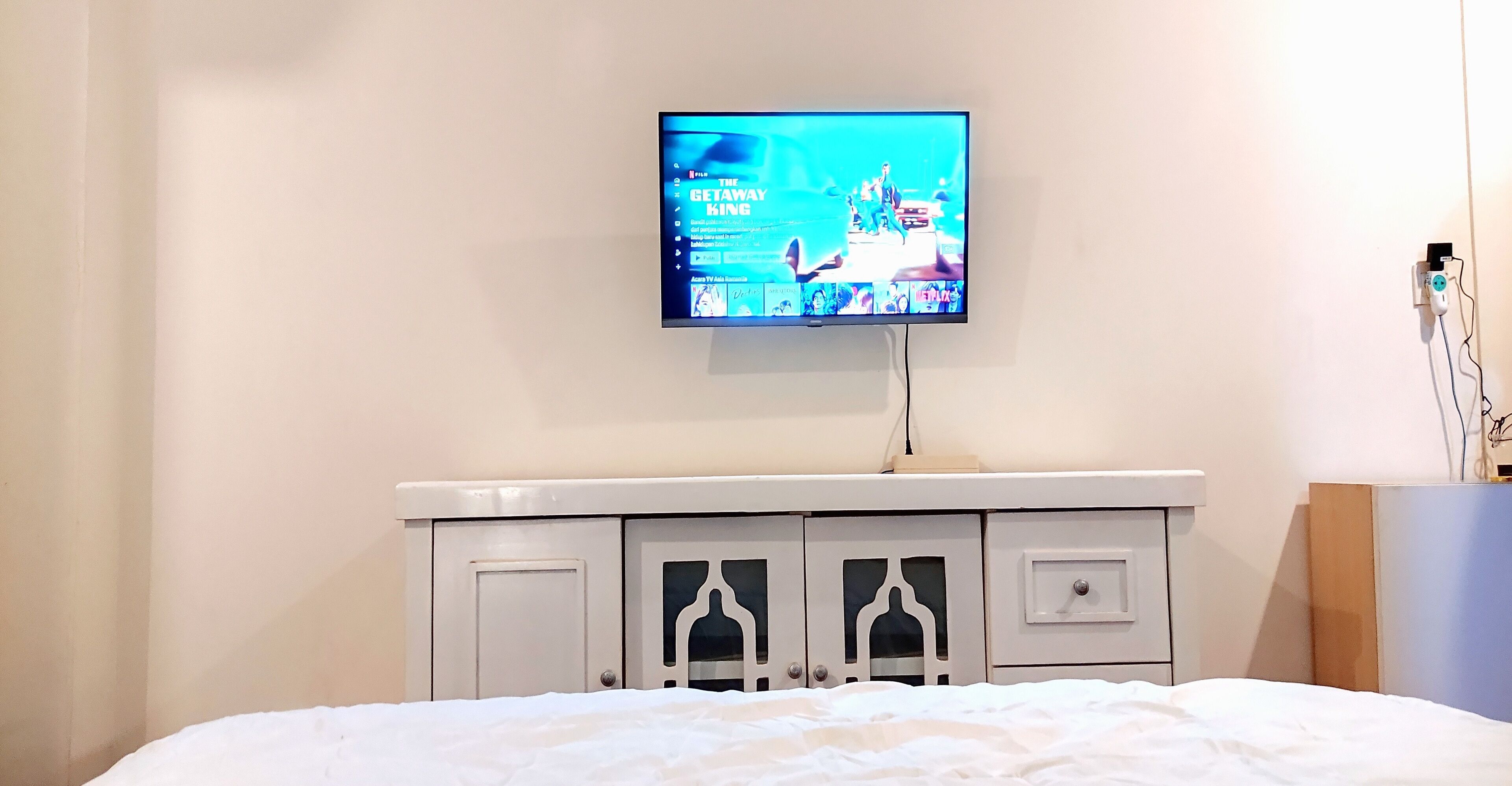A bedroom with a bed, a TV mounted on the wall, and a white dresser with drawers and a cabinet.