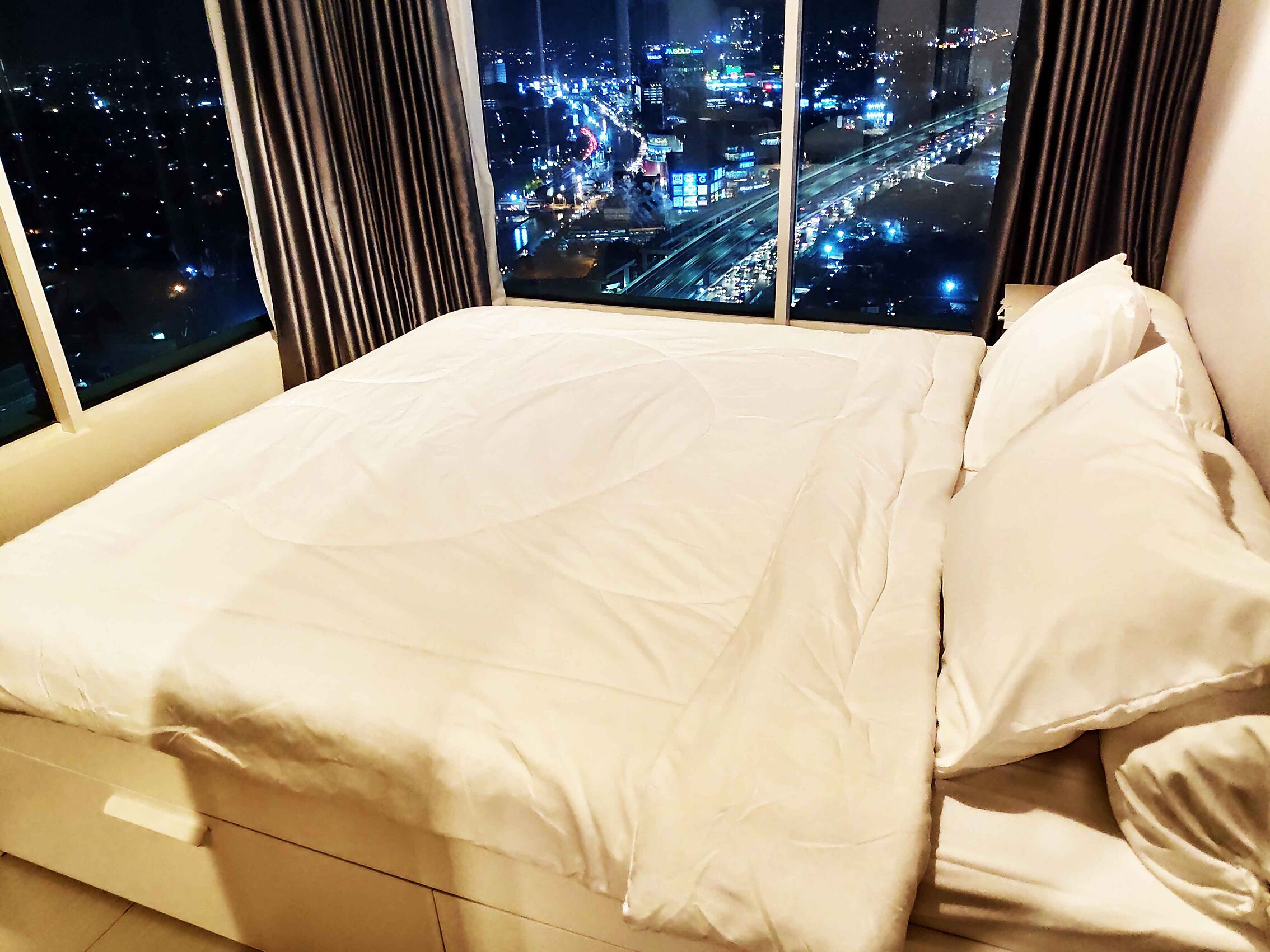 Una cama con ropa de cama blanca en una habitación con ventanas grandes que ofrecen vistas nocturnas de la ciudad.
