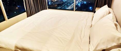 Una cama con ropa de cama blanca en una habitación con ventanas grandes que ofrecen vistas nocturnas de la ciudad.