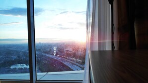 Et værelse med et stort vindue der vender ud mod en byudsigt i solnedgang.