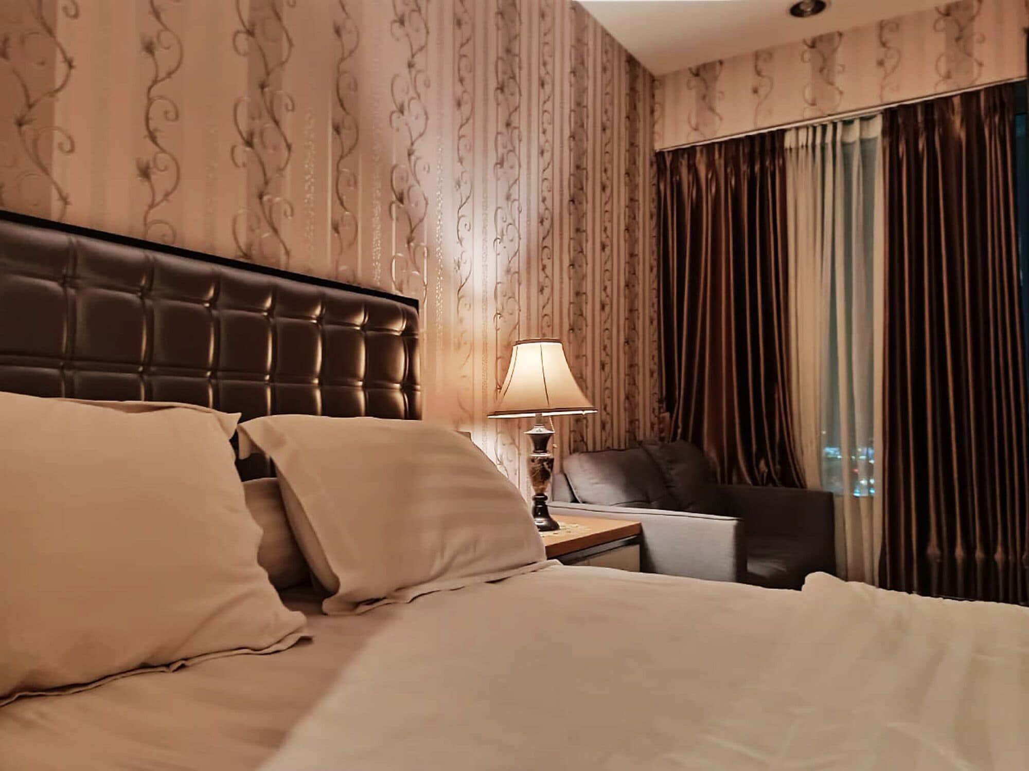 A hotel room with a tufted headboard, a bed with beige linens, a nightstand with a lamp, and a view of the city through large curtains.