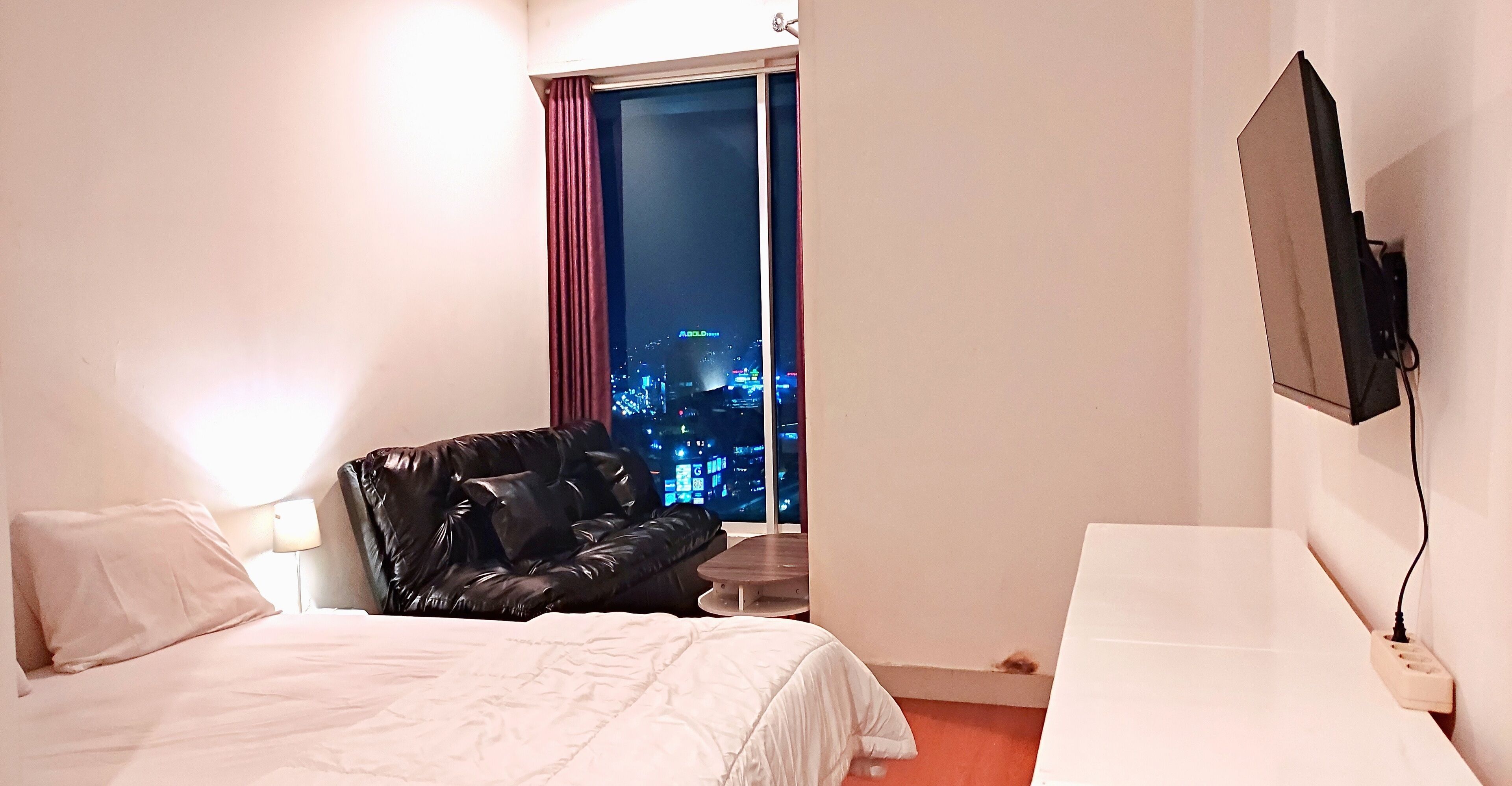 Habitación de hotel con cama, un sillón de cuero negro, una mesita pequeña, un televisor montado en la pared y una ventana con vista nocturna a la ciudad.