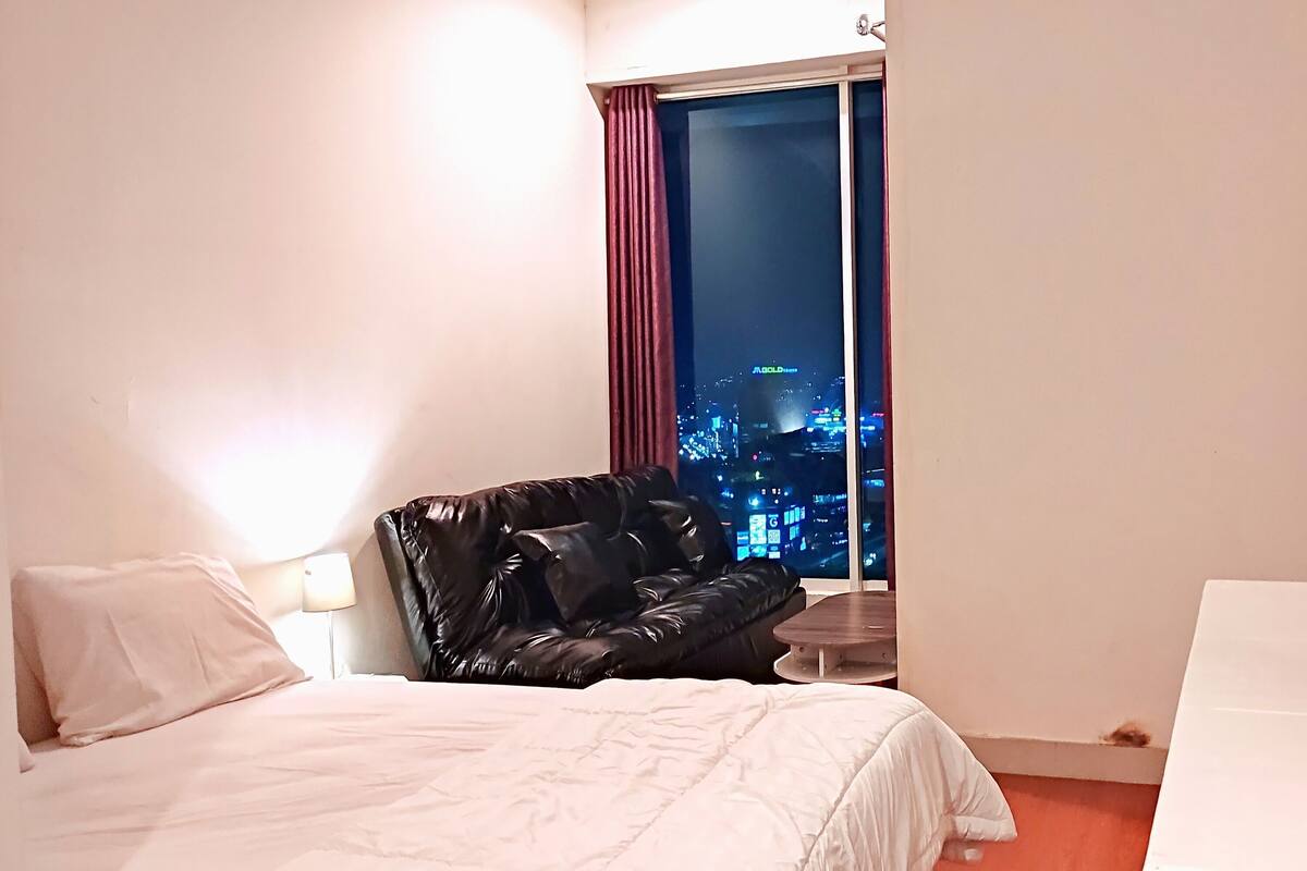 Ett hotellrum med en säng, en svart läderfåtölj, ett litet bord, en tv som är monterad på väggen och ett fönster med utsikt över stadens skyline på natten.